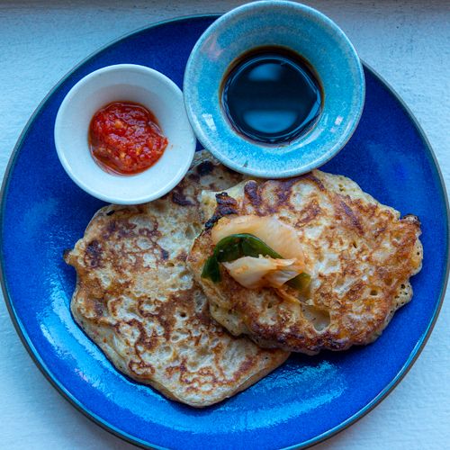 Two kimchi pancakes on a blue plate, with a small bowl of dipping sauce and a small bowl of hot sauce.